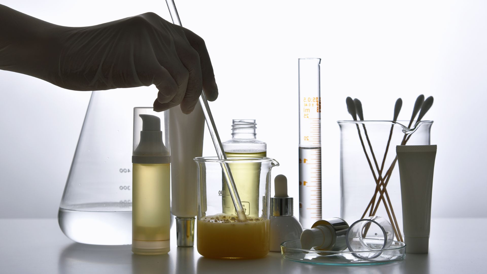 A scientist wearing gloves mixing ingredients in a beaker, surrounded by skincare product packaging and lab tools.